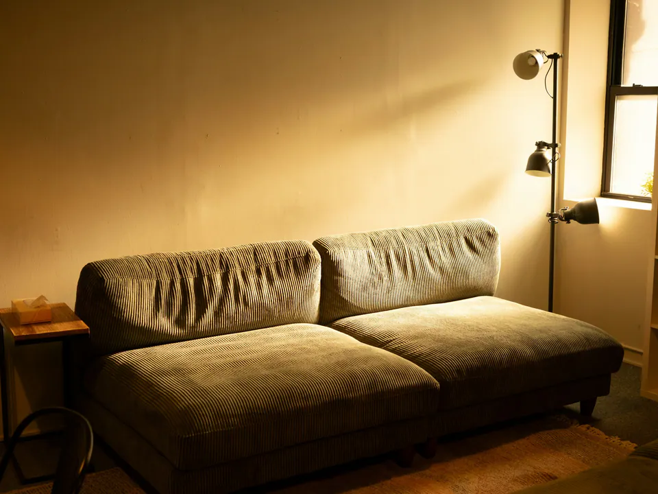 A sofa washed in warm light from a wall lamp at dusk, beside a shelf and a window