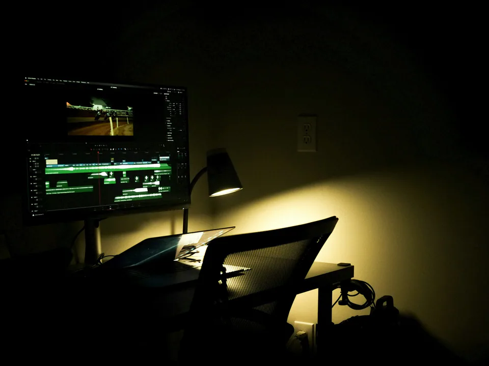 An edit desk in a dark room, a monitor showing a timeline and a desk lamp pooling warm light