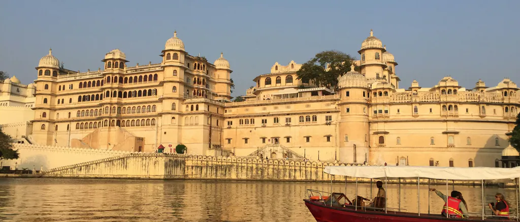 The City Palace in Udaipur seen from Lake Pichola in warm evening light, with a red boat on the water