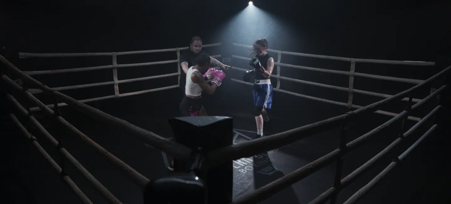 Film still from Piroba: two boxers and a referee in a ring under a single overhead spotlight, seen past a corner post