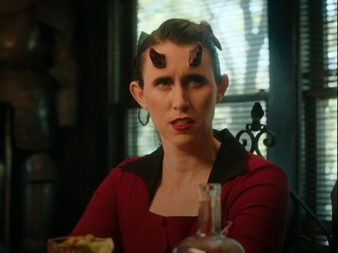 Film still from The Morningstars: a woman with small horns and red lipstick frowns across a dinner table