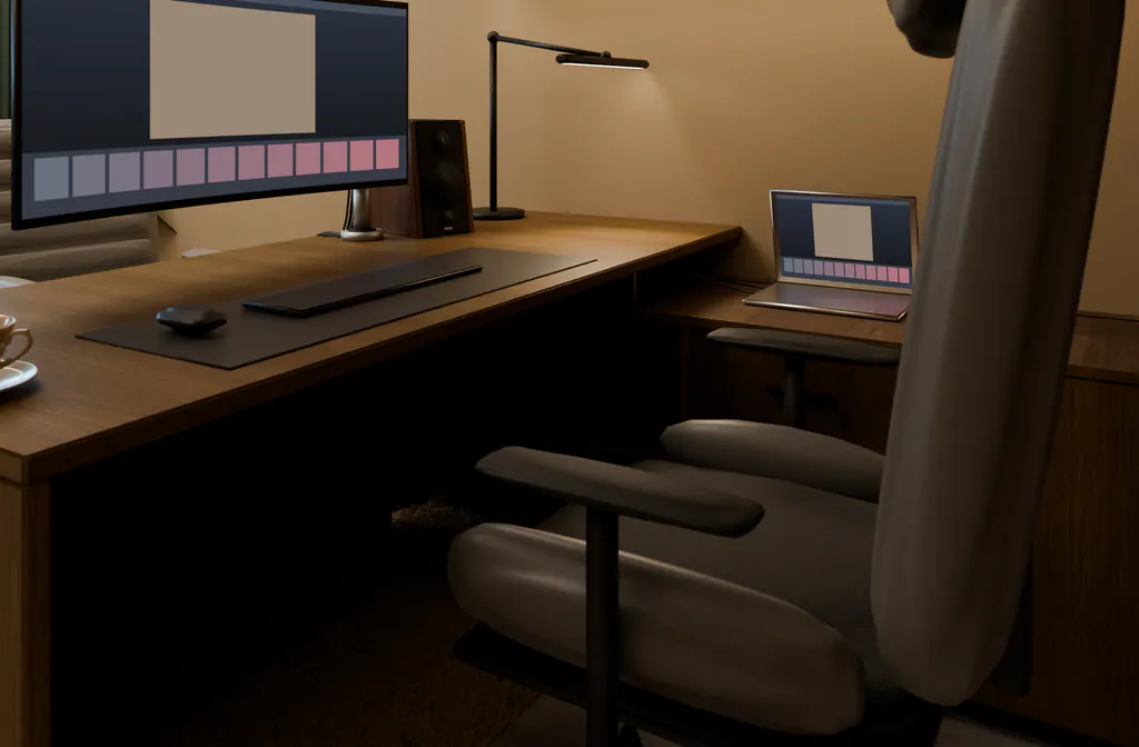Low, close view behind an office chair: a curved monitor, a desk lamp throwing a pool of warm light, and deep shadow under the desk.